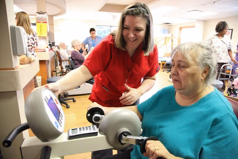 A healthcare worker in a red uniform assists an elderly woman using an exercise machine in a bright room with other elderly people and staff in the background.