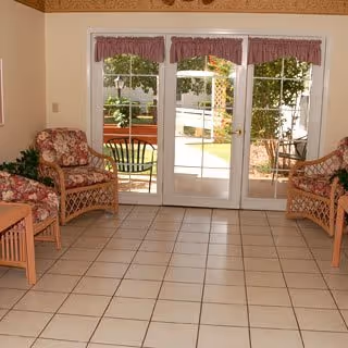 Sunlit common sitting area with wicker floral-upholstered chairs facing glass doors that open to a garden patio.