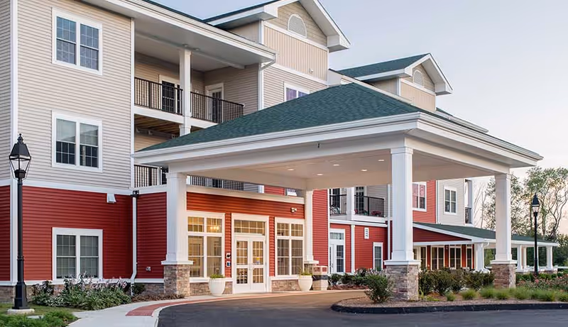 Exterior view of a senior living facility building with a covered entrance supported by white columns. The building has beige and red siding with multiple windows and balconies. There are landscaped bushes and a paved driveway in front.