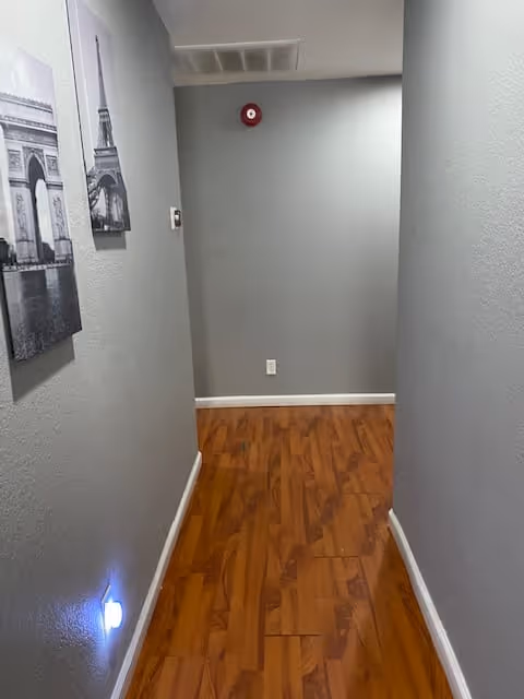 Narrow interior hallway with gray walls, wood-look flooring, wall art on the left, and a red fire alarm on the far wall.