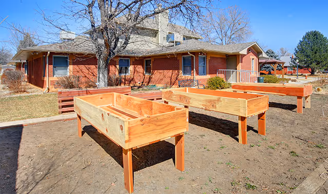 Outdoor garden area with three raised wooden planter boxes on a dirt patch in front of a red brick building under a clear blue sky.