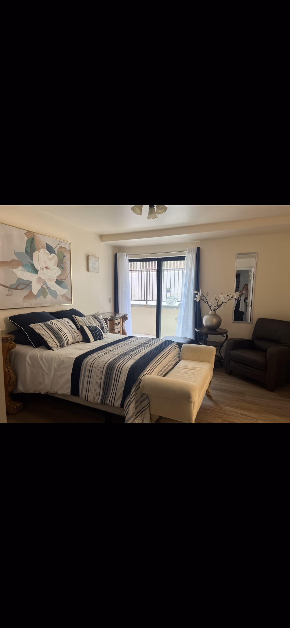 A cozy bedroom with a neatly made bed featuring striped bedding in navy and white. There is a cushioned bench at the foot of the bed, a dark armchair to the right, and a small round table with a decorative vase and flowers. A large floral painting hangs on the wall above the bed, and a tall mirror is mounted on the wall near the armchair. A sliding glass door with white curtains leads to an outdoor area.