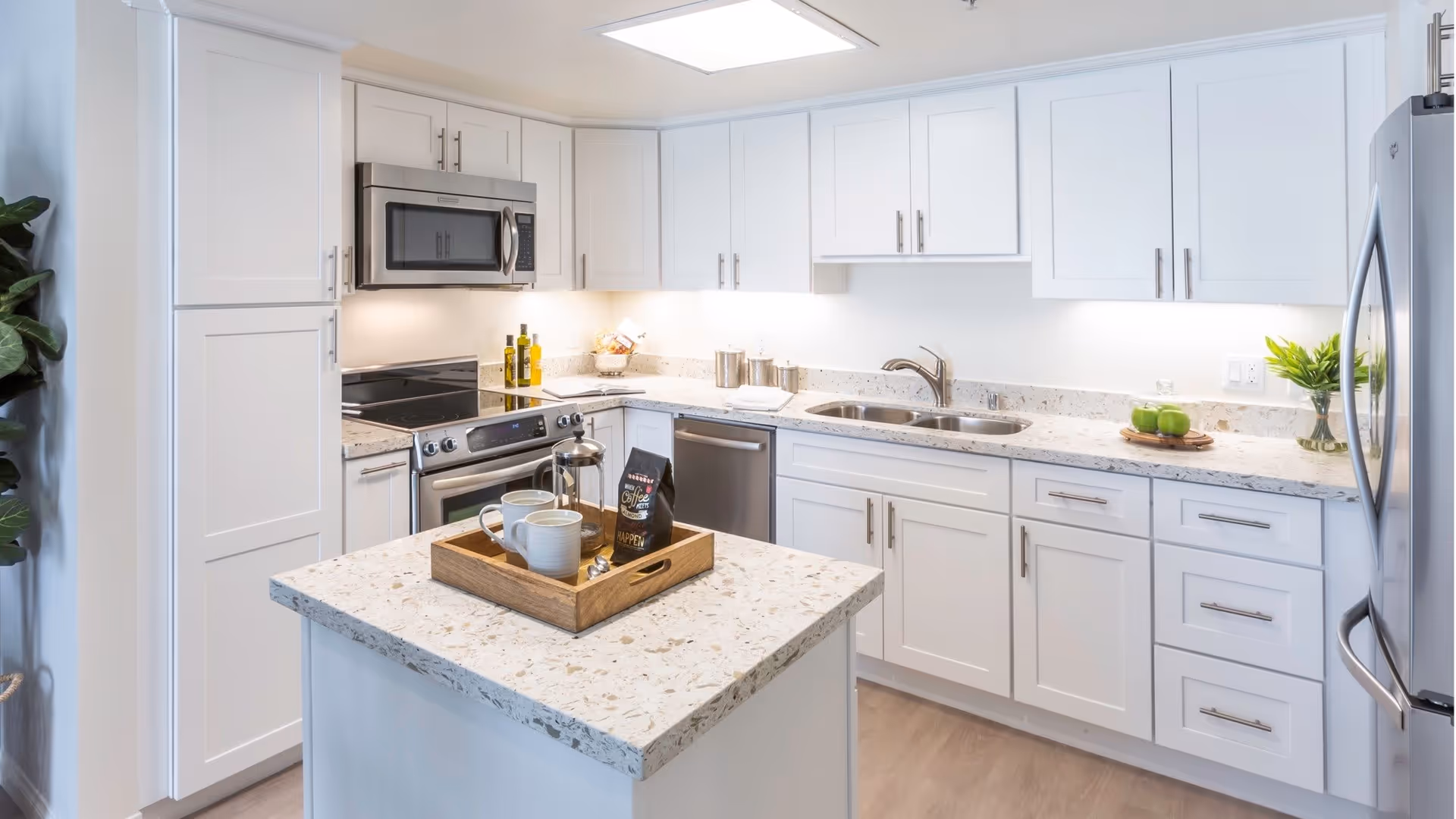 Bright modern kitchen with white cabinets, a central island with a wooden tray holding a French press and two mugs, stainless steel appliances including a microwave, stove, dishwasher, and refrigerator, and a countertop decorated with green apples and a small plant.