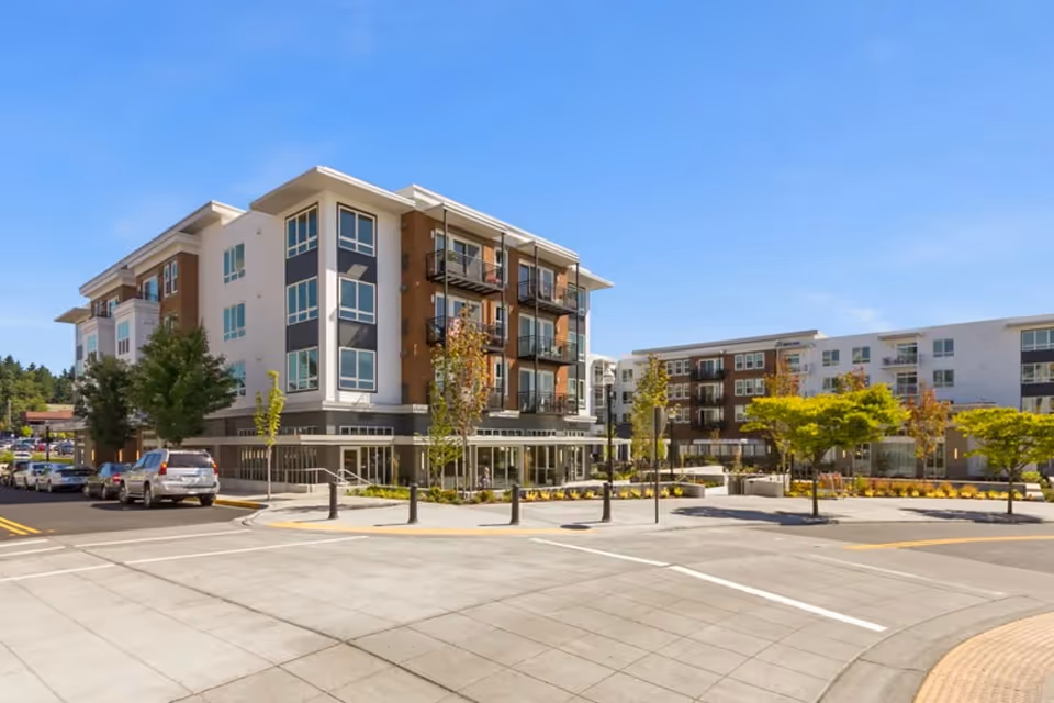 Modern four-story apartment building with balconies, street parking, and a landscaped plaza under a clear blue sky.