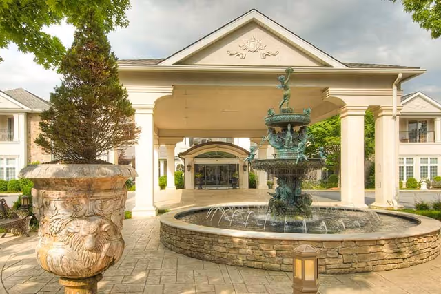 Entrance of a senior living building with a covered porte-cochère, a central stone fountain, and a large decorative planter.