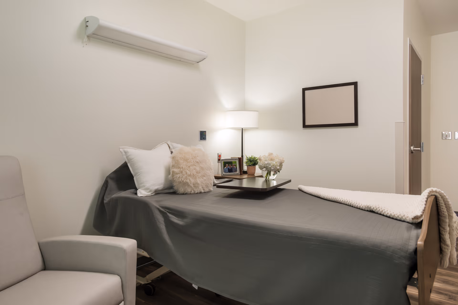 A neatly made single bed with gray bedding and white pillows in a small, clean room. A bedside table holds a lamp, a small plant, a photo frame, and a vase with flowers. There is a light gray armchair to the left and a blank picture frame on the wall. The room has light-colored walls and wooden flooring.
