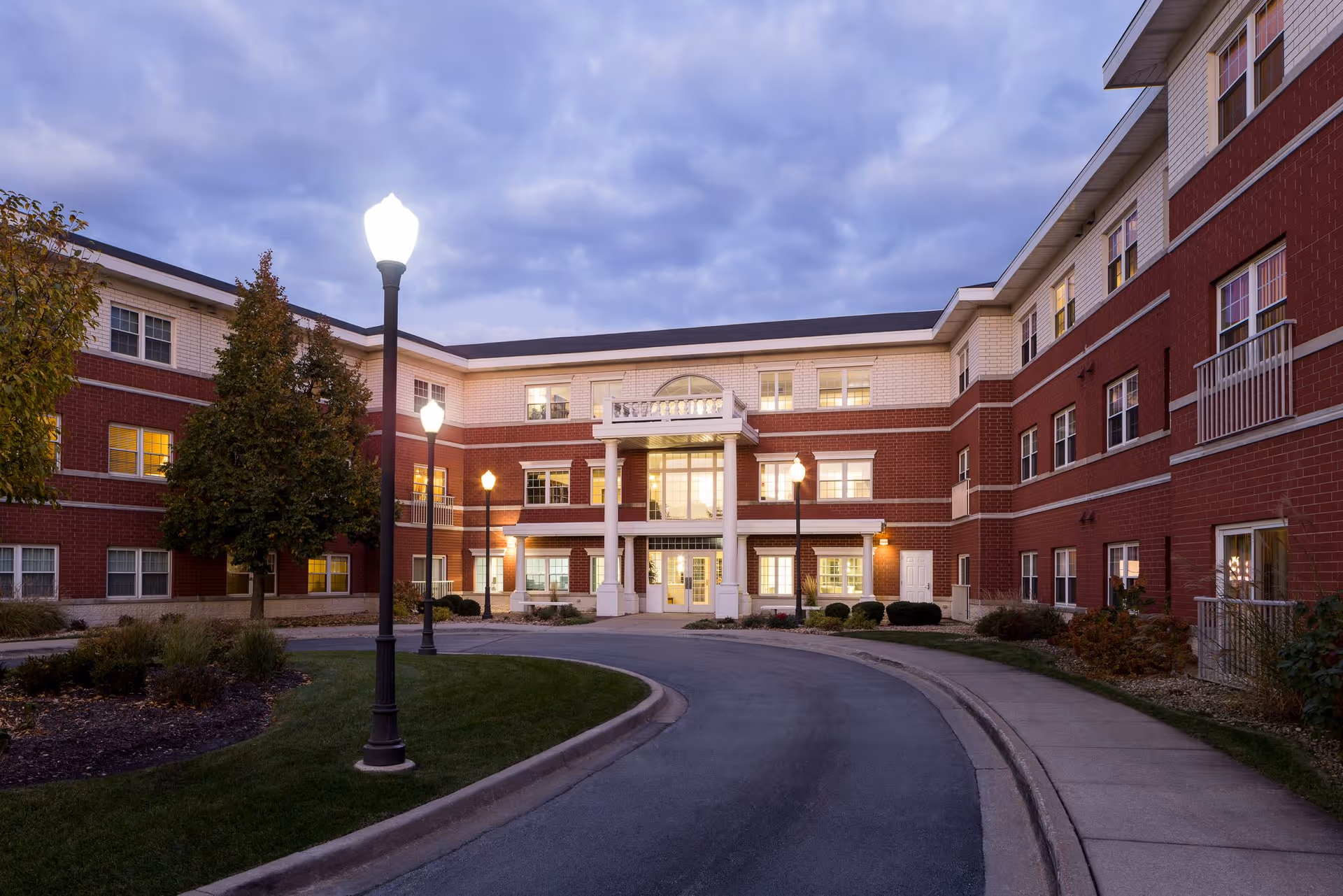 Exterior view of a three-story senior living facility building named Hanover Place during dusk, with illuminated windows and street lamps along a curved driveway leading to the entrance.