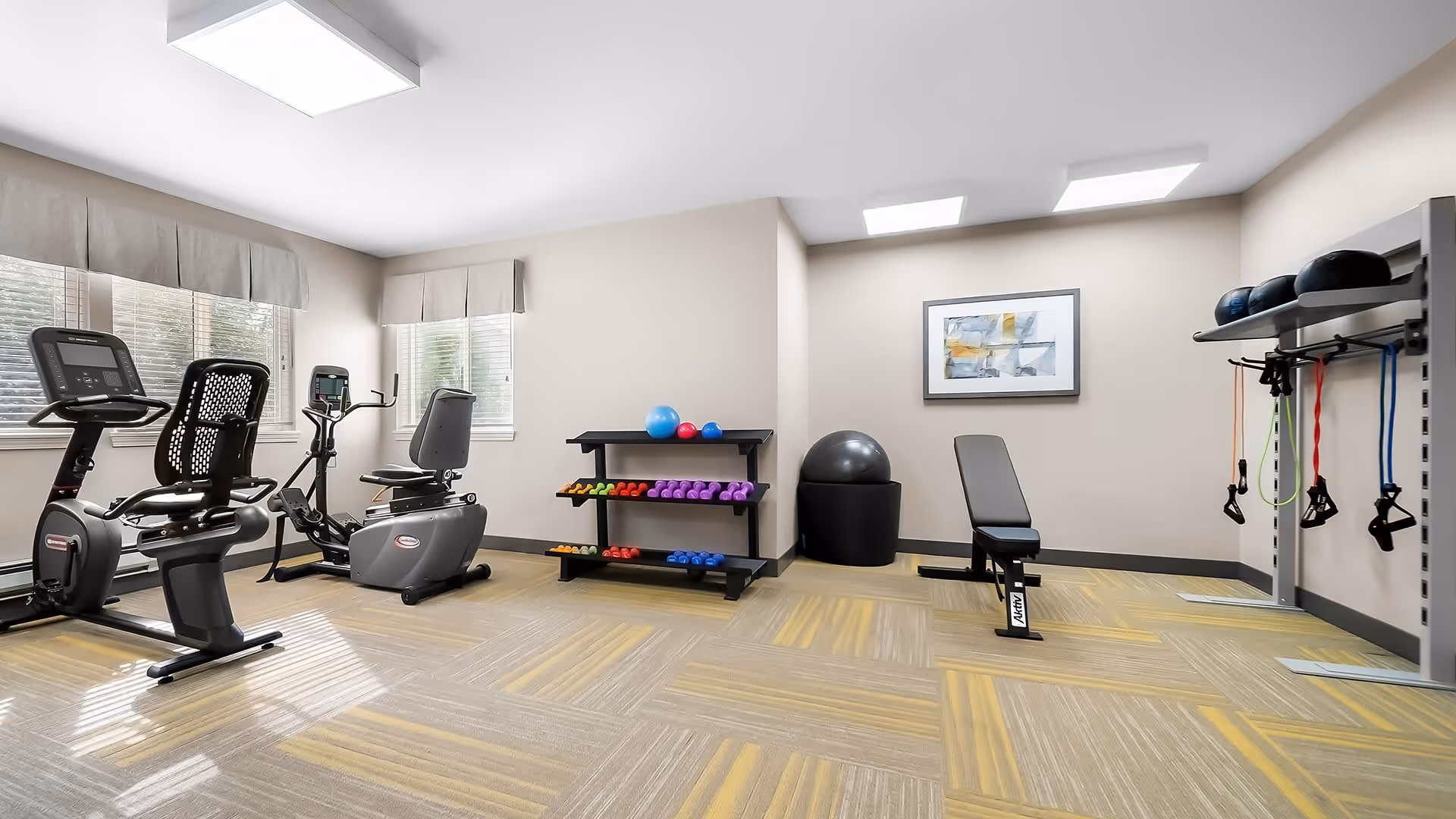A bright and clean exercise room with two recumbent stationary bikes, a rack of colorful dumbbells, exercise balls, resistance bands hanging on a wall rack, and a workout bench. The room has large windows with valances, light-colored walls, and patterned carpet flooring.