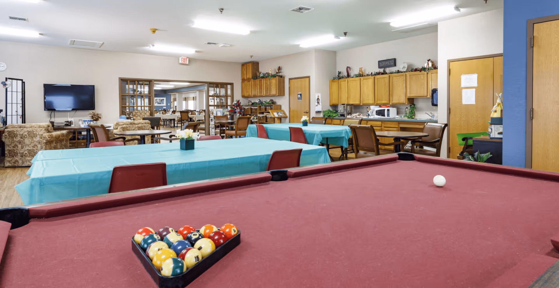 Interior view of a senior living common room with a pool table in the foreground, tables with teal tablecloths, seating and a kitchenette in the background.