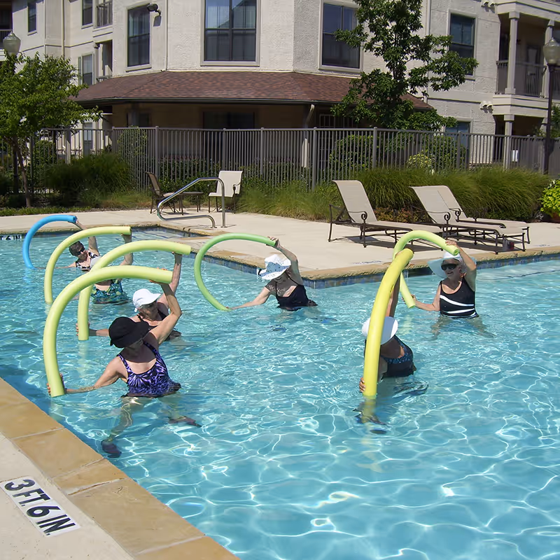 Several people wearing sun hats doing water aerobics with foam pool noodles in an outdoor swimming pool beside a multi-story residential building.