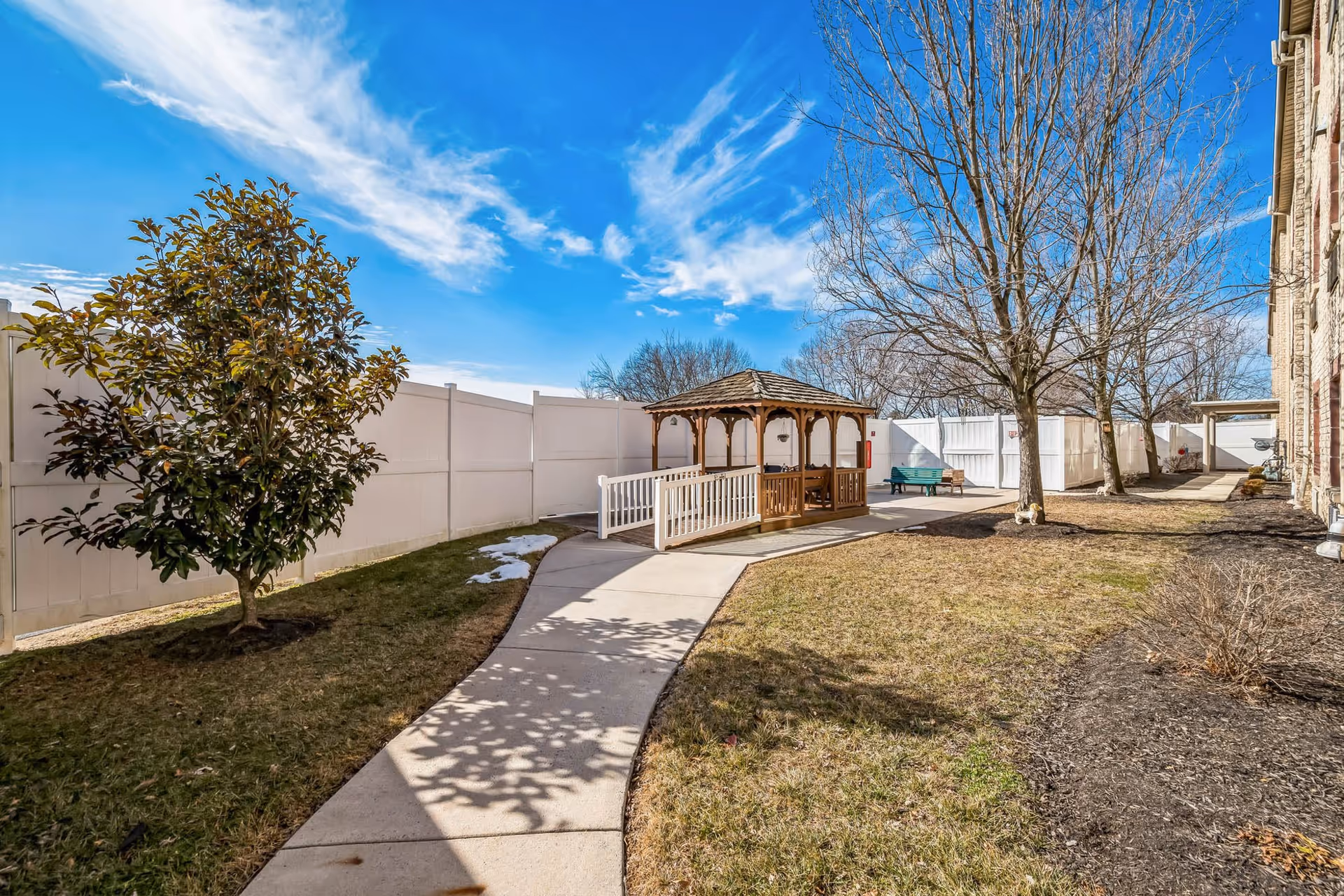Outdoor area at Greenfield Senior Living at Graysonview featuring a paved walkway leading to a wooden gazebo with seating. The area is surrounded by a white fence, with trees and grass on either side under a partly cloudy blue sky.