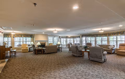 Spacious senior living facility common area with multiple seating arrangements including armchairs and sofas, large windows with valances allowing natural light, a fireplace, and carpeted floor.