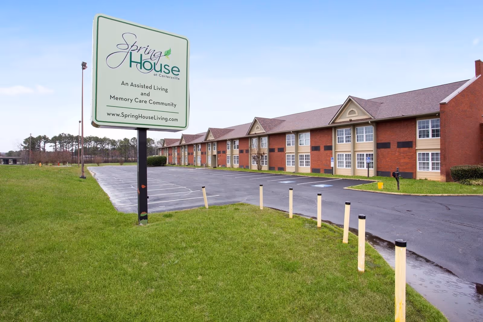 Exterior view of Springhouse at Cartersville, an assisted living and memory care community, showing a large sign with the facility's name and website, a parking lot, and a two-story brick building with multiple windows.