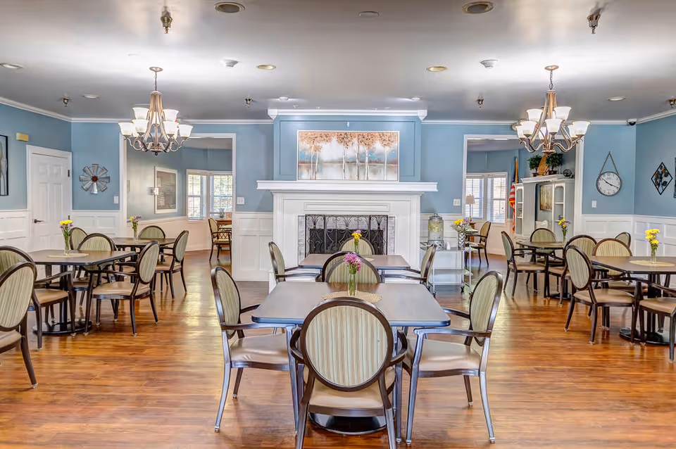 Bright communal dining room with multiple tables and chairs, hardwood floors, chandeliers, and a central fireplace under soft blue walls.