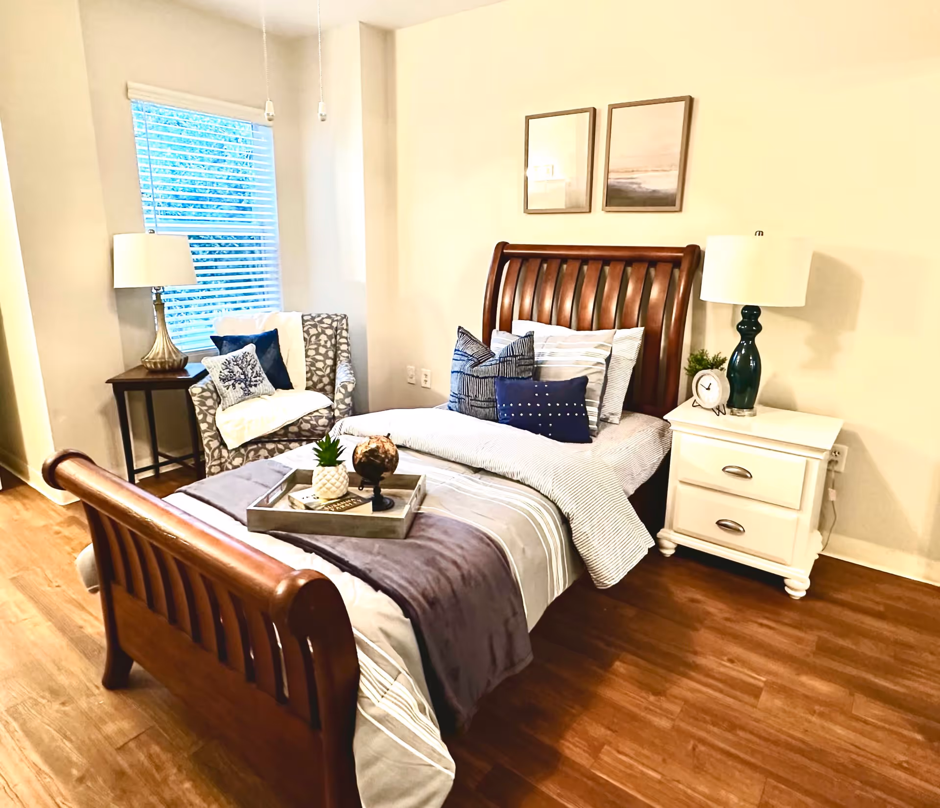 A cozy bedroom with a wooden sleigh bed made up with striped bedding and multiple decorative pillows. A tray with a small globe and a pineapple decor sits on the bed. To the right of the bed is a white nightstand with a green lamp, a small clock, and a plant. To the left, there is a patterned armchair with a white throw blanket and blue pillows next to a window with blinds and a table lamp on a small side table. The room has wooden flooring and light-colored walls with two framed pictures hanging above the bed.