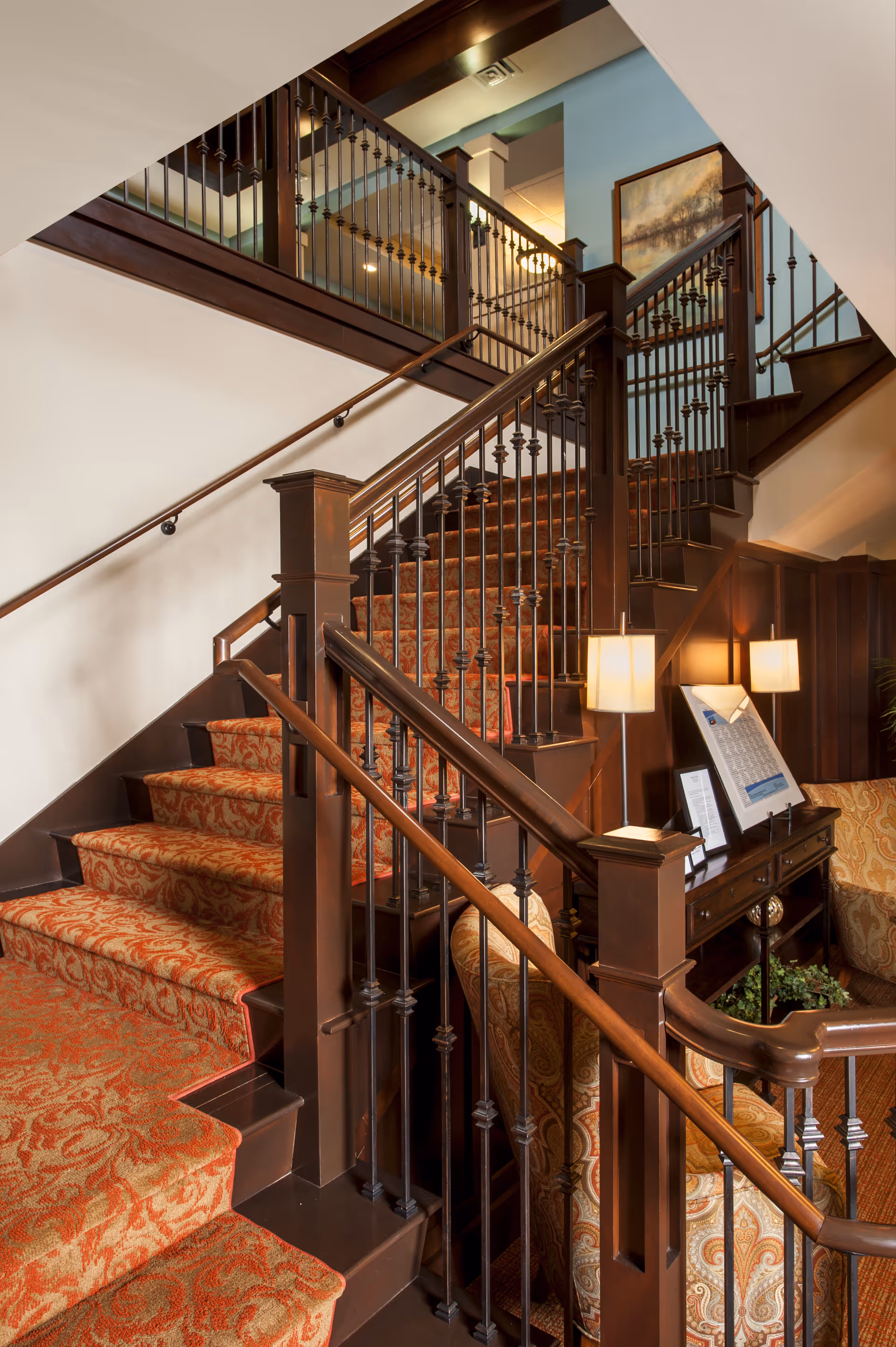 Interior view of a senior living facility showing a carpeted staircase with ornate wooden railings and iron balusters. The stairs are covered with a red and gold patterned carpet. There is a small seating area with a patterned armchair and a wooden console table with lamps and framed pictures. The walls are painted light blue and white, and a framed painting hangs on the wall above the stairs.