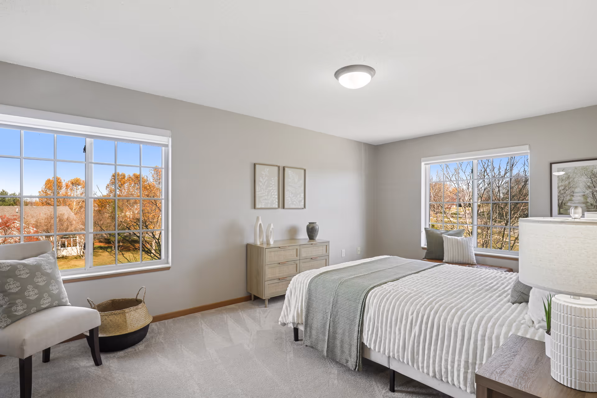 A bright and airy bedroom with two large windows showing autumn trees outside. The room features a neatly made bed with white and gray bedding, a wooden dresser with decorative vases, a cushioned chair with a patterned pillow, a basket on the floor, and a bedside table with a lamp and small plant.