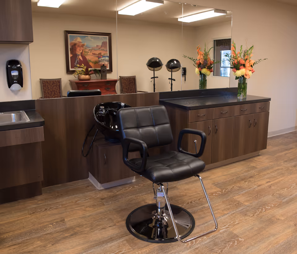 A salon area inside Ashton Medical Lodge featuring a black salon chair in front of a black wash basin. The room has wooden cabinets with a black countertop, a large mirror, a vase with orange and yellow flowers, and a painting of a woman wearing a hat on the wall. The floor is wooden, and there is a hand sanitizer dispenser mounted on the wall near a small sink.