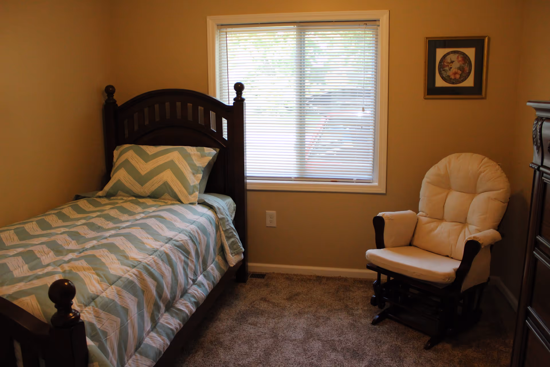 A small bedroom with a single wooden bed featuring a green and white chevron patterned bedspread and pillow. Next to the bed is a cushioned beige rocking chair. A window with closed blinds is centered on the wall behind the bed and chair. A framed floral picture hangs on the wall above the chair. The floor is carpeted in a neutral tone.