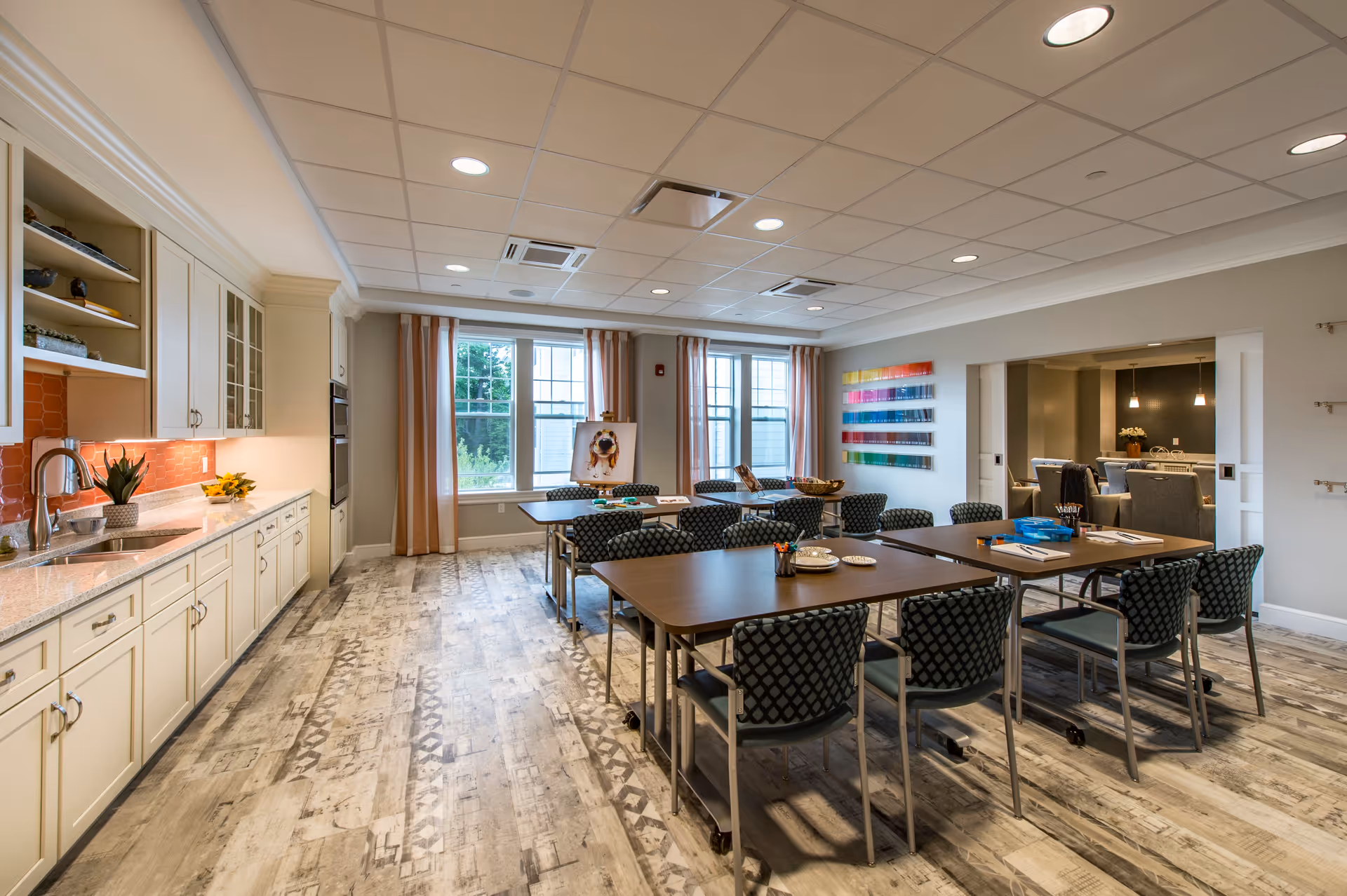 Communal dining/activity room featuring tables and patterned chairs, a kitchenette along the left wall, and large windows with curtains.
