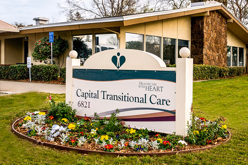 Outdoor view of the Capital Transitional Care facility sign surrounded by a flower bed with colorful flowers and greenery, with the building and windows visible in the background.
