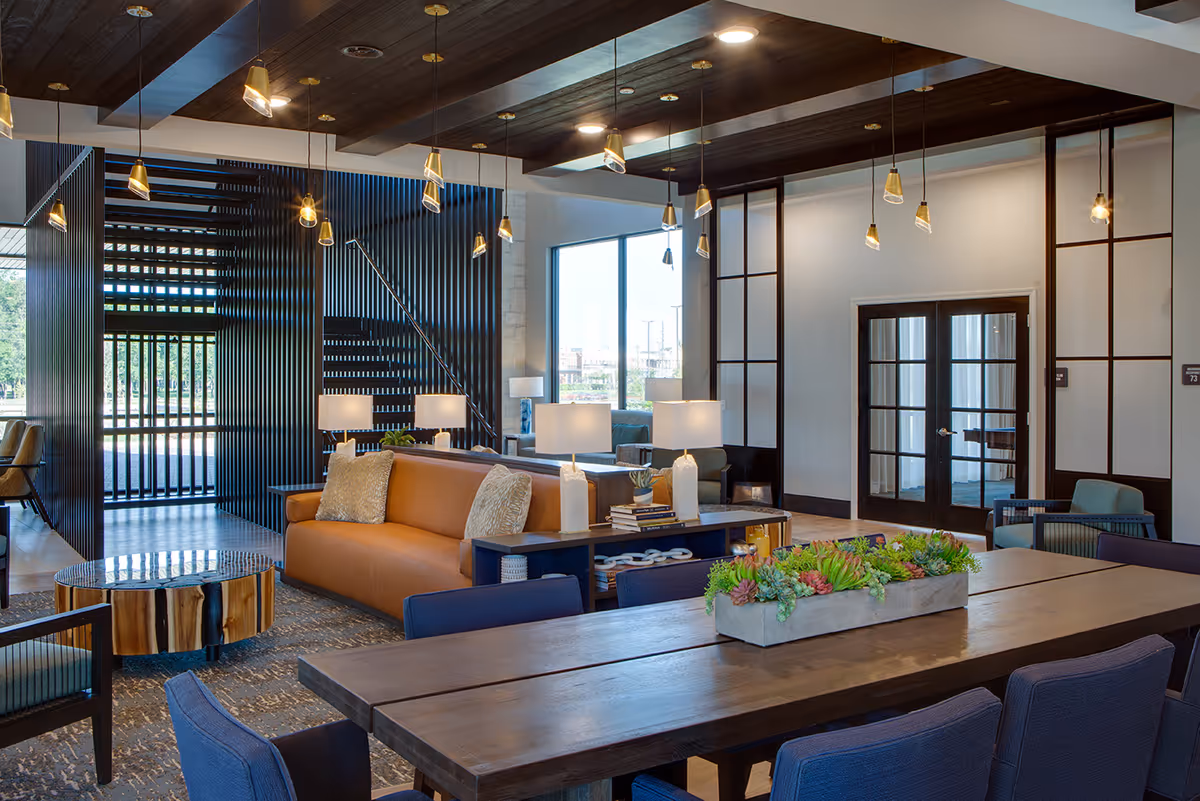 Modern senior living facility common area with a large wooden table decorated with a rectangular planter of succulents, surrounded by blue upholstered chairs. Behind the table is a brown leather sofa with two pillows, a wooden coffee table with a natural wood finish, and several table lamps. The space features a dark wood ceiling with multiple hanging pendant lights, large windows letting in natural light, and a staircase with black vertical slats.