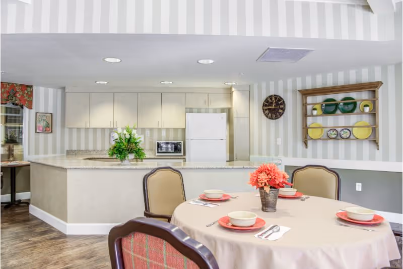 A dining area with a round table set for four with bowls, plates, and utensils. A vase with pink flowers is in the center of the table. Behind the table is a kitchen with white cabinets, a refrigerator, a microwave, and a countertop. The walls have light-colored vertical stripes, and a wooden shelf with decorative plates is mounted on the wall. A clock is also visible on the wall.