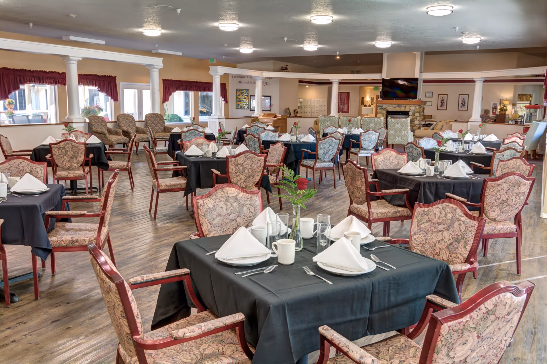 A spacious dining room in a senior living facility with multiple tables covered in black tablecloths, each set with white napkins, plates, cups, and silverware. The chairs have floral upholstery and wooden frames. The room features large windows with red curtains, wooden flooring, white columns, and a stone fireplace with a TV mounted above it.