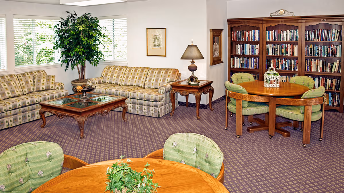 A cozy living room area with two patterned sofas, a wooden coffee table with glass inserts, a side table with a lamp, and a large potted plant near windows with blinds. There are two wooden bookshelves filled with books and a round wooden table surrounded by four cushioned chairs with green upholstery. The carpet is patterned in purple tones.