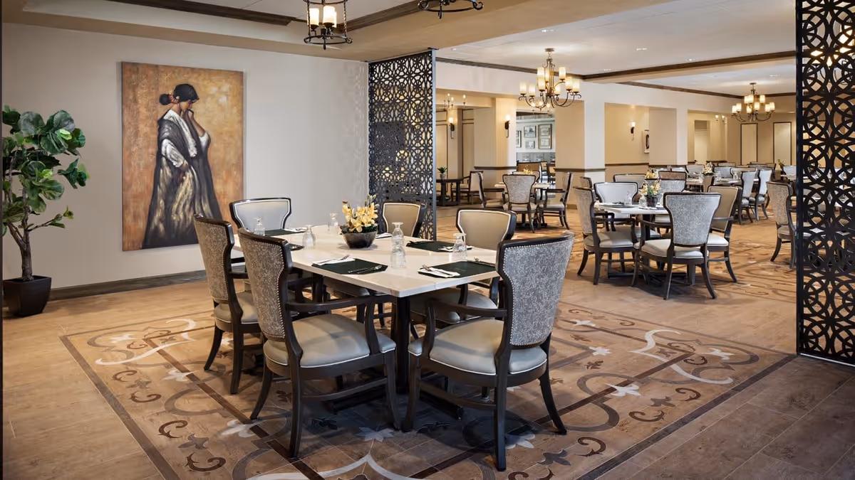 Elegant communal dining room with several set tables and upholstered chairs, decorative room dividers, and chandeliers.