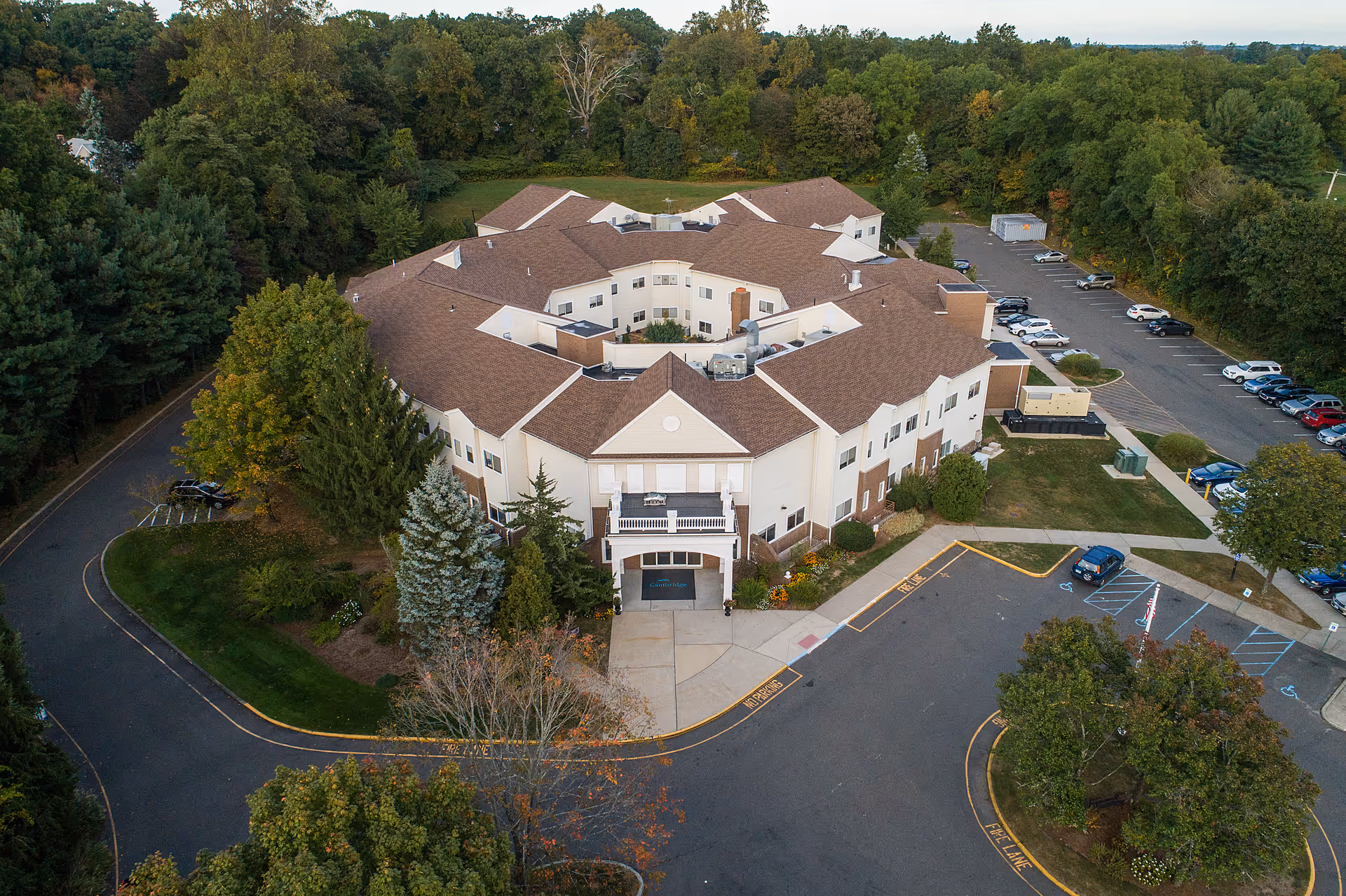 Aerial view of Cambridge Health and Rehabilitation Center, a large two-story building with a brown roof surrounded by trees and parking lots. The building is situated in a green area with a driveway and several parked cars visible.