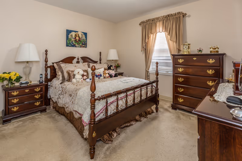 A cozy bedroom with a wooden bed frame adorned with stuffed animals and decorative pillows. The room features matching wooden nightstands with lamps, a wooden dresser, a window with beige curtains, and beige carpeting.