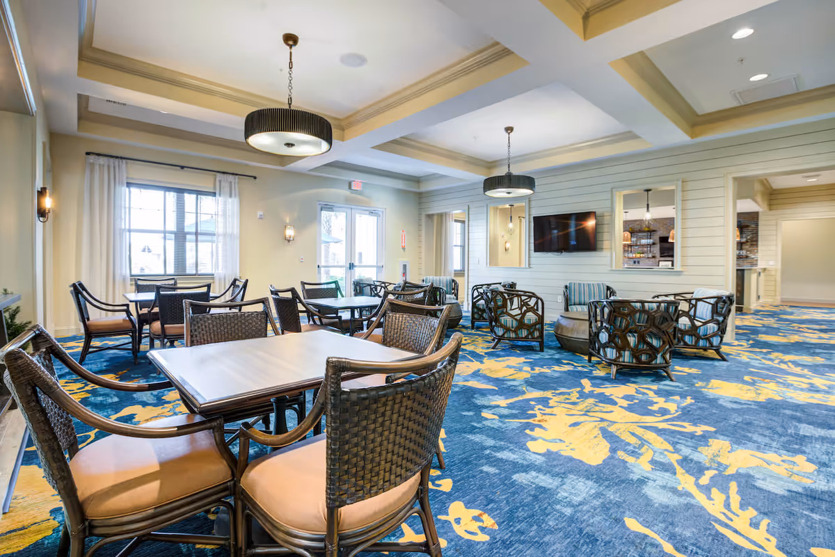 A bright and spacious common area in a senior living facility featuring multiple tables with wicker chairs and a seating area with patterned armchairs and a round ottoman. The room has large windows with sheer curtains, modern pendant lights hanging from a coffered ceiling, and a wall-mounted flat-screen TV. The carpet is blue with yellow floral patterns, and there are doorways leading to other rooms.