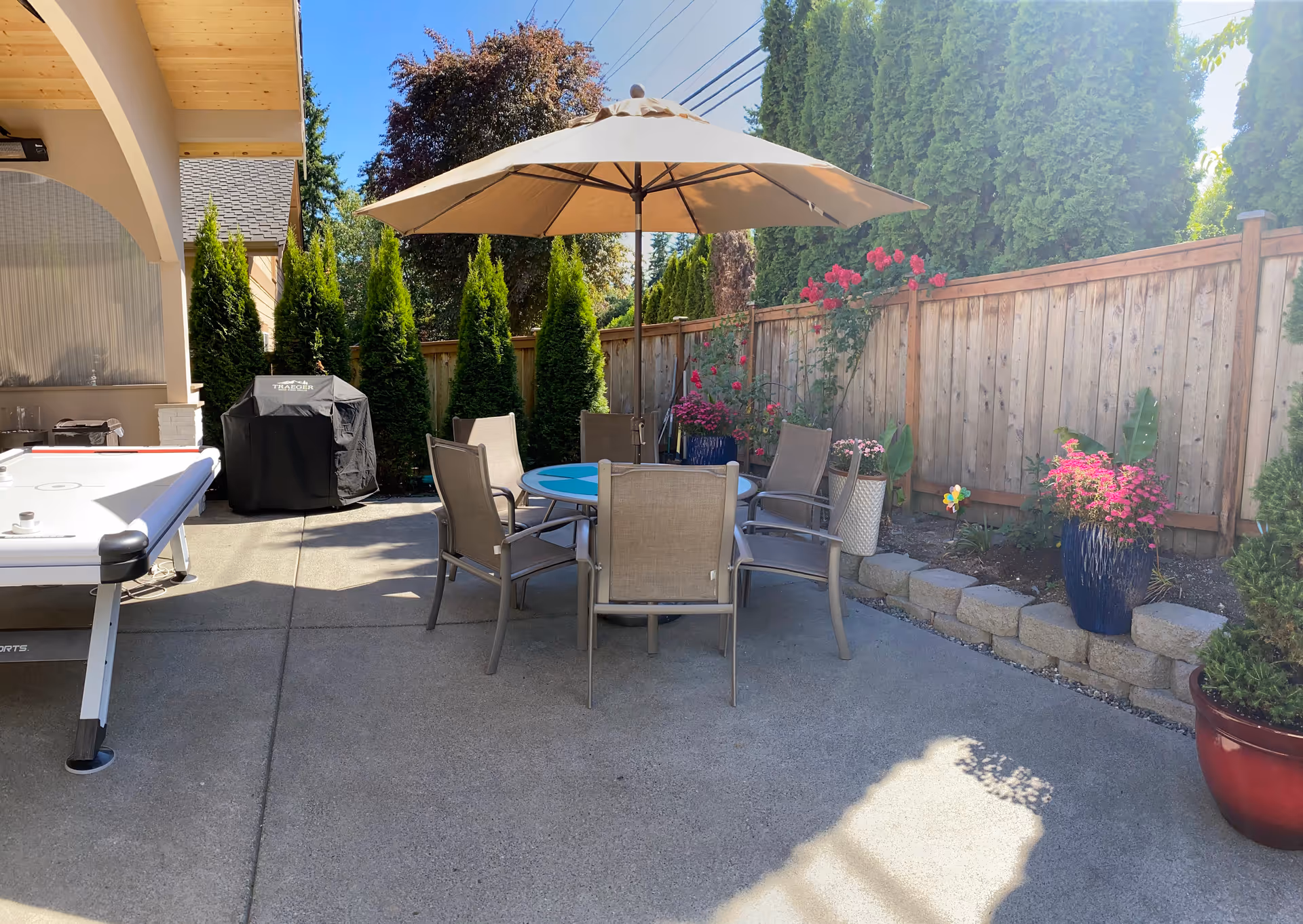 Outdoor patio area with a round table and six chairs under a large beige umbrella. There are potted plants and flowers along a wooden fence, a covered grill, and an air hockey table on the concrete floor. Tall green trees and shrubs provide privacy around the space.