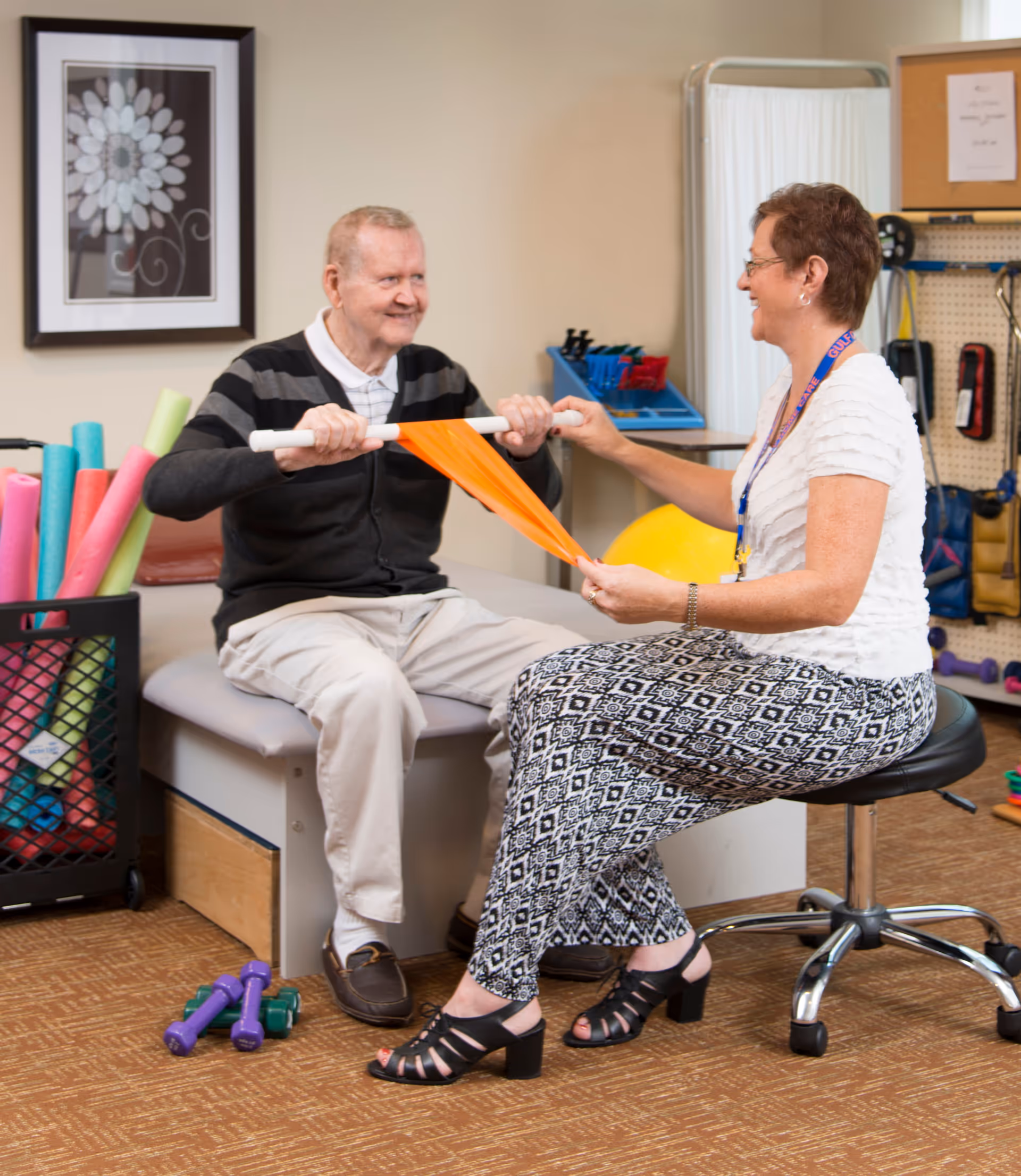 An elderly man and a woman engage in a physical therapy exercise using an orange resistance band in a therapy room. The man is seated on a padded bench holding a white bar attached to the band, while the woman, seated on a rolling stool, assists him. The room contains various therapy equipment including colorful foam rollers, dumbbells, and other exercise tools.