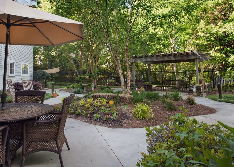 Outdoor garden area at Sunrise on Providence featuring a round table with chairs under a large beige umbrella, a paved walkway, various plants and flowers, trees providing shade, and a wooden pergola with seating underneath.