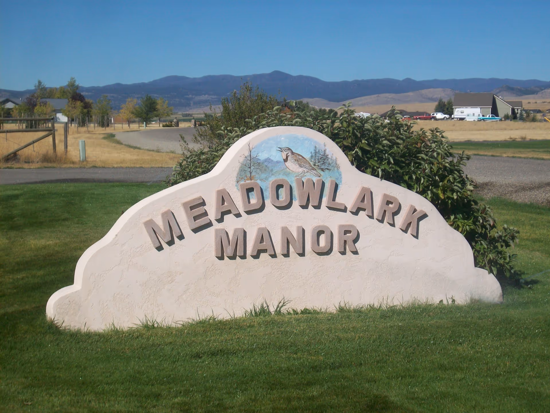 A large stone sign with the words 'Meadowlark Manor' carved into it, set on a grassy area with bushes behind it. In the background, there are fields, a few buildings, and mountains under a clear blue sky.