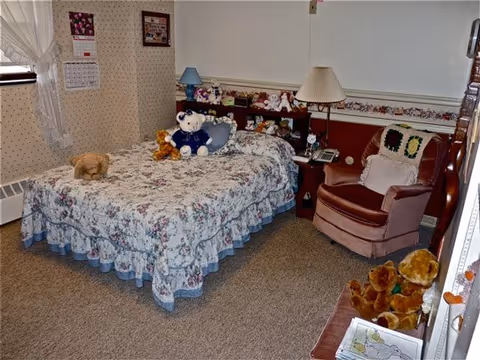 Cozy bedroom with a floral-covered single bed topped with stuffed animals, a pink armchair, and a bedside table with a lamp.