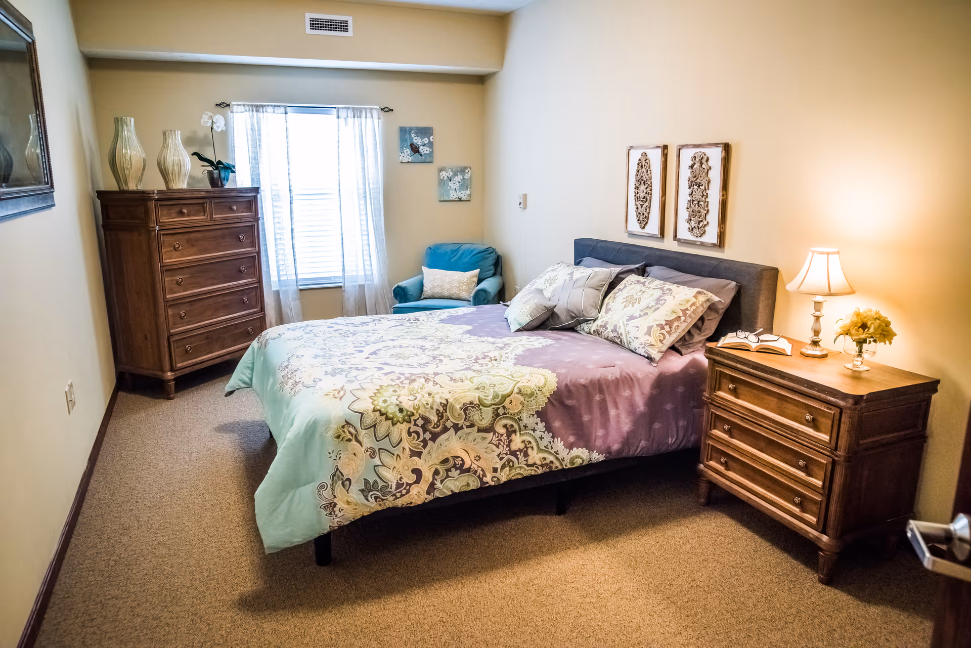 A cozy bedroom with a bed covered in a patterned comforter and multiple pillows. There is a wooden nightstand with a lamp, a book, and a small flower arrangement on the right side of the bed. On the left side of the room, there is a tall wooden dresser with decorative vases and a plant on top. A blue armchair is placed near a window with sheer white curtains. Two framed decorative wall hangings are above the bed.