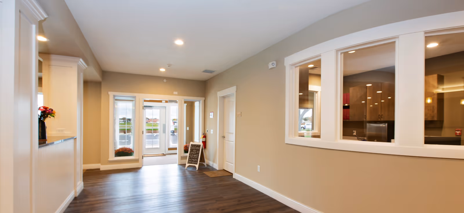 Interior view of a senior living facility hallway with wood flooring, beige walls, and white trim. There are large windows on the right side looking into another room, a small chalkboard sign near the glass entrance doors, and a fire extinguisher mounted on the wall.
