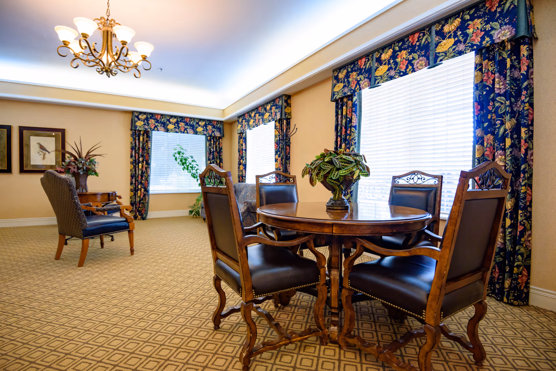 A well-lit common area with a round wooden table surrounded by four upholstered chairs. The room features large windows with floral-patterned curtains, a chandelier hanging from the ceiling, framed bird artwork on the wall, and additional seating with a patterned armchair and side table with a plant.