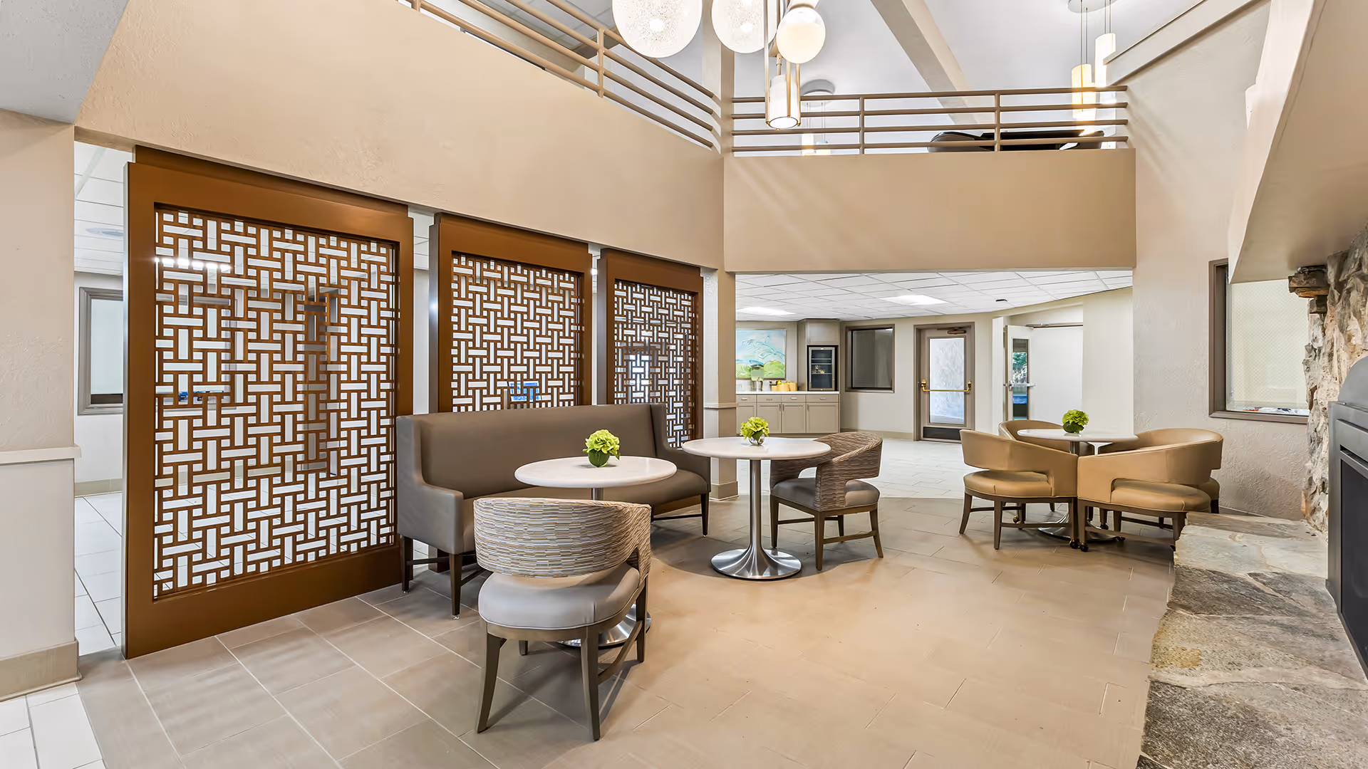 A bright and spacious common area in a senior living facility featuring modern round tables with chairs and a bench, decorative wooden lattice panels on the wall, a stone fireplace on the right, and a high ceiling with hanging light fixtures.