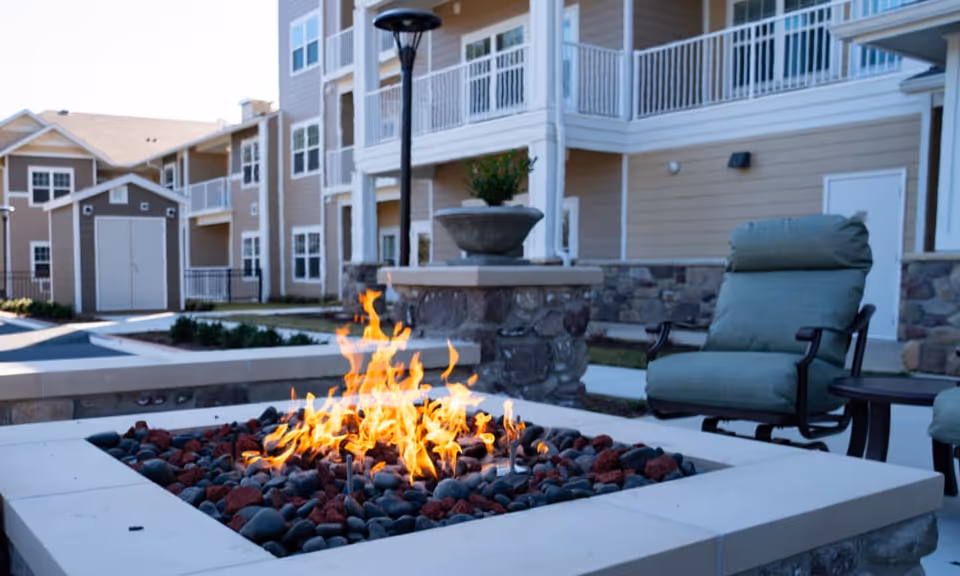 Outdoor seating area at a senior living facility featuring a modern fire pit with flames surrounded by smooth black and red stones, cushioned chairs, a small table, and a multi-story beige building with balconies in the background.