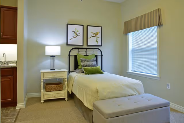 A neatly made single bed with green and patterned pillows in a small bedroom. Next to the bed is a white nightstand with a lamp and a wicker basket on the lower shelf. Two framed bird illustrations hang on the wall above the bed. A window with blinds and a valance is on the right wall, and part of a kitchenette with a sink and wooden cabinets is visible on the left.