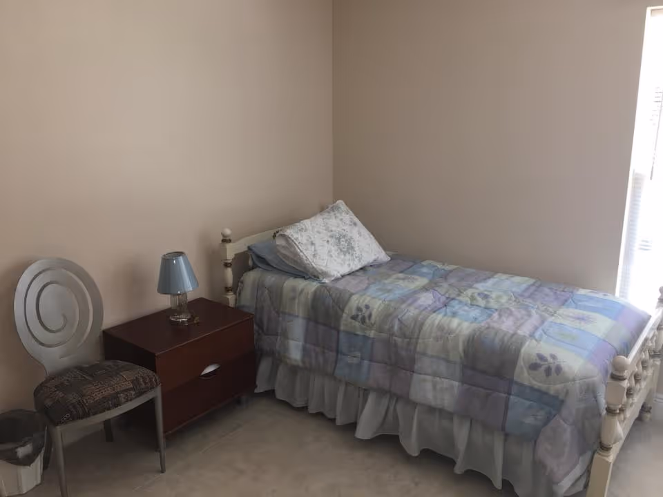 A small bedroom with a single bed covered with a pastel-colored patchwork quilt and a floral pillow. Next to the bed is a wooden nightstand with a small blue lamp on it. A chair with a patterned cushion is placed beside the nightstand. The room has beige walls and a window with blinds partially open, letting in natural light.