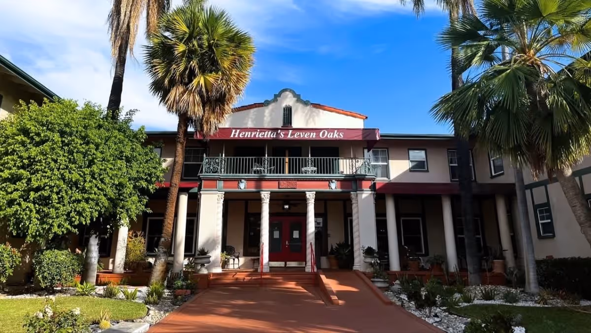 Front exterior view of Henrietta's Leven Oaks by SCH, a two-story building with a red awning displaying the facility's name. The entrance features white columns, a ramp, and stairs, surrounded by palm trees and landscaped greenery under a blue sky.