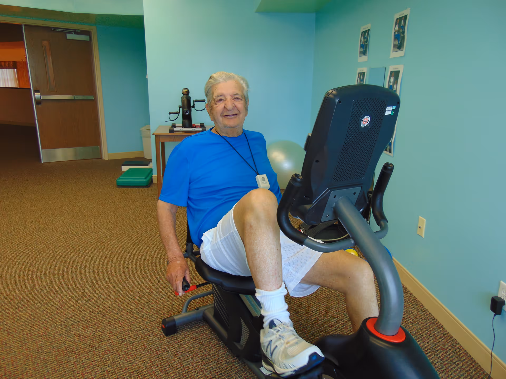 An elderly man wearing a blue shirt and white shorts is sitting on a recumbent exercise bike in a room with light blue walls and brown carpet. He is smiling and appears to be exercising. There is exercise equipment and framed pictures on the wall behind him.