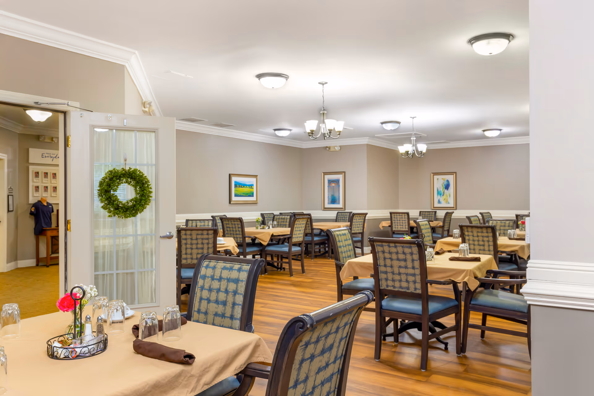 Well-lit dining room with multiple tables set with beige tablecloths and chairs, hardwood floors, framed artwork, and a wreath on a glass door.