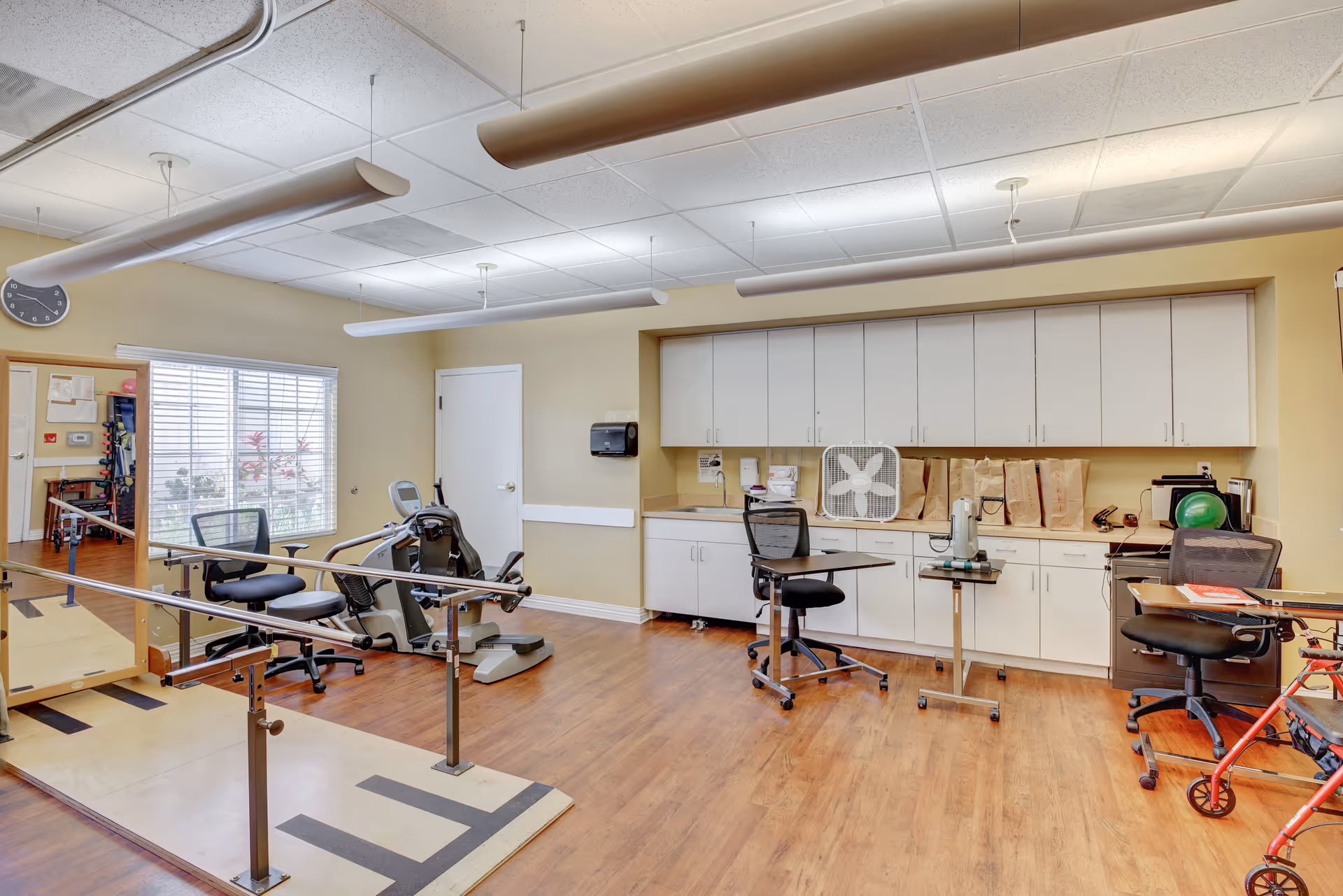 A rehabilitation or physical therapy room with parallel bars for walking exercises, exercise equipment including a recumbent bike, office chairs, a walker, and cabinets along the wall. The room has wood flooring, a window with blinds, and fluorescent ceiling lights.