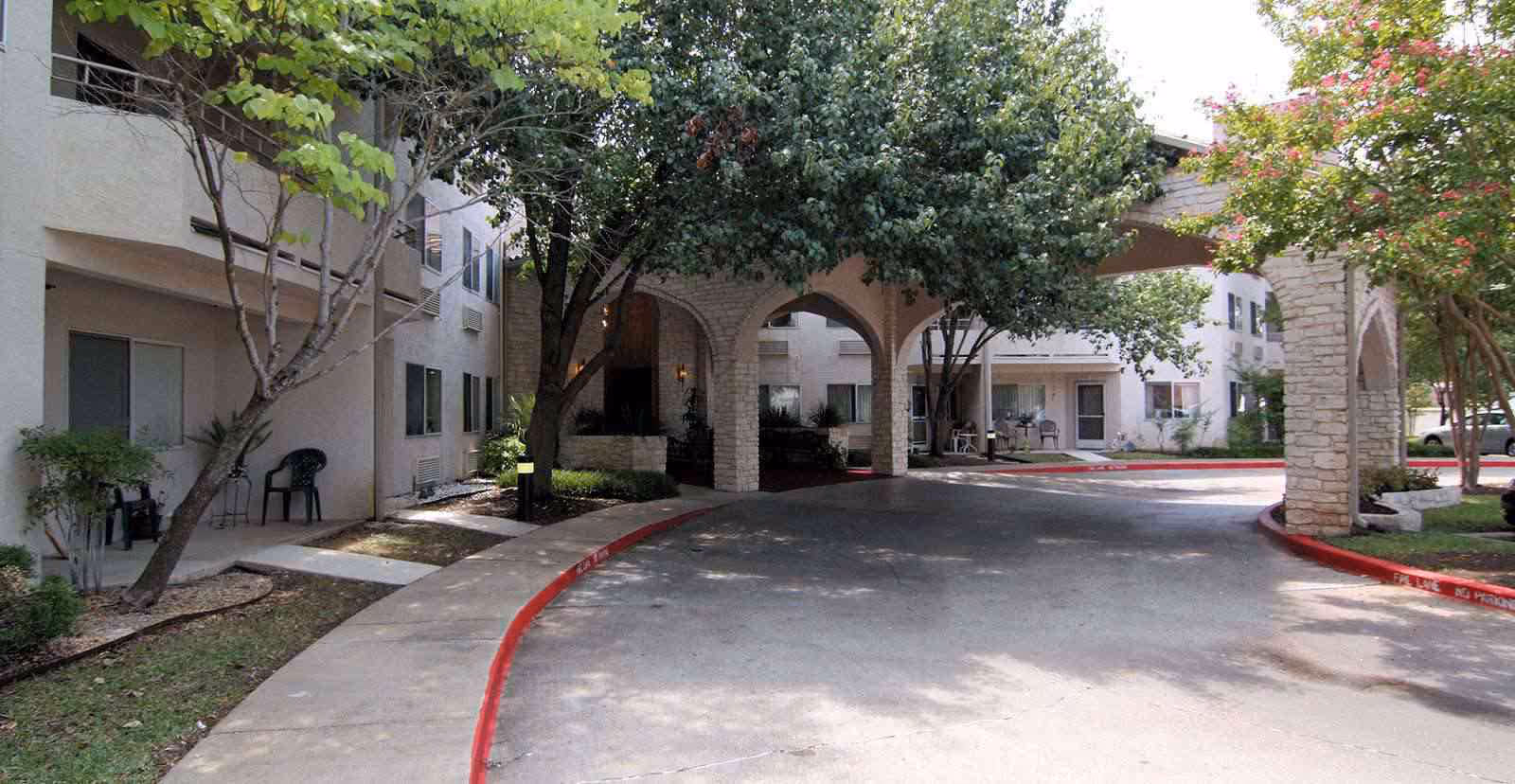 Exterior view of Solstice Senior Living at Austin showing a driveway with a covered entrance supported by stone arches, surrounded by trees and shrubs, with a multi-story building in the background featuring windows and patio doors.